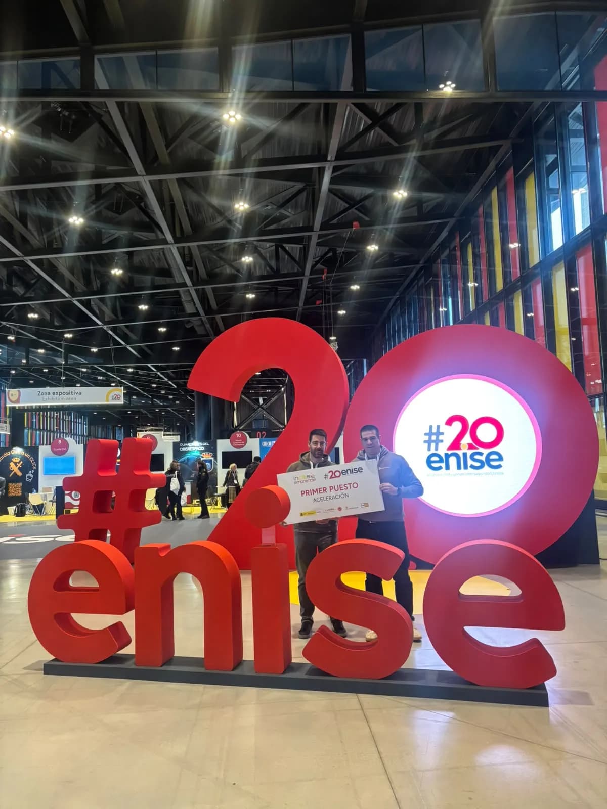 Two members of the Montevive team holding the INCIBE Emprende first place Acceleration board, in front of the giant #20ENISE letters